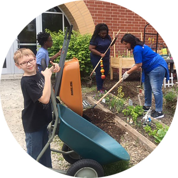 Kids working in a small space garden in front of a school. Two are using wheelbarrows and two are using long-handled garden tools.