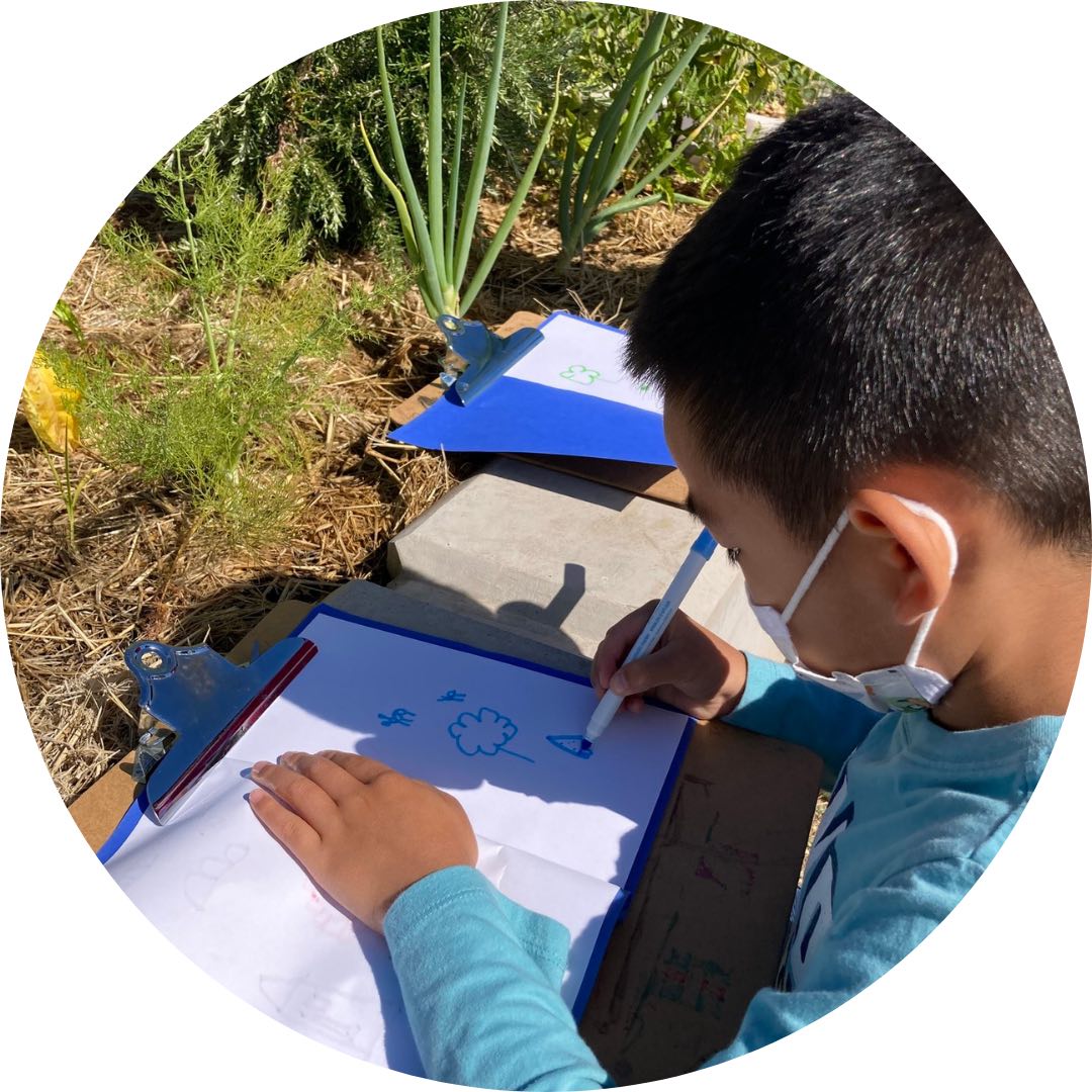 A child holding a pen, writing on a clipboard. They are seated in a garden.
