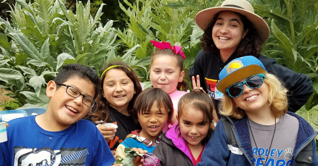 3 Six elementary-age smiling, happy kids with a young adult. In the background are garden crops.