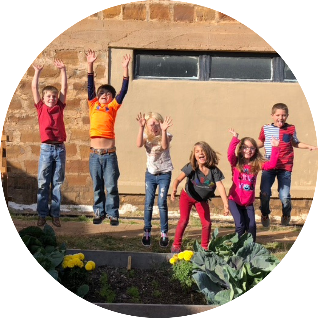 Children jumping up and crouching down, about to jump. There is garden produce in front of them and a building behind them.