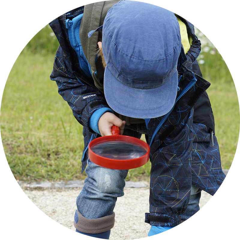 kid with blue hat outside bent over looking at the ground through magnifying glass